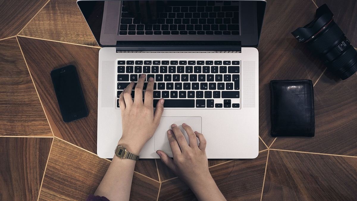 Hands typing on a laptop with a smartphone beside it on a wooden desk